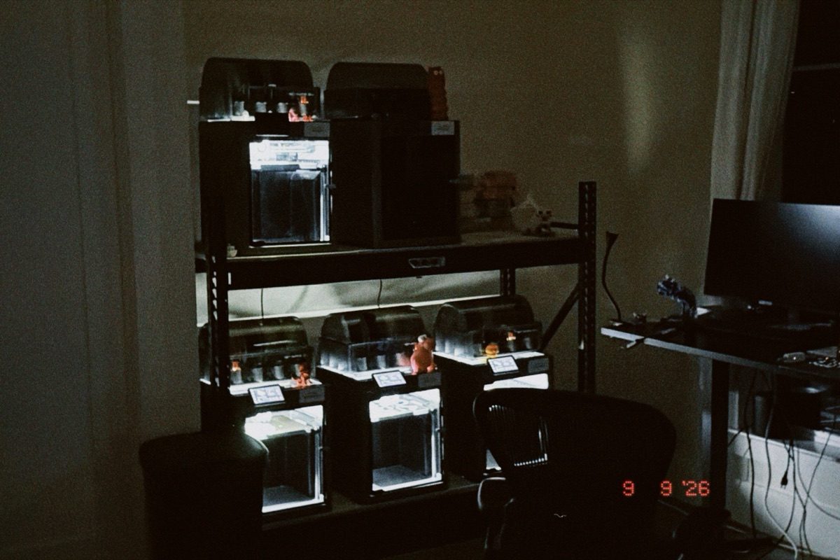 A steel shelving unit holding six enclosed 3D printers glowing in a dark room, small toys on top of each