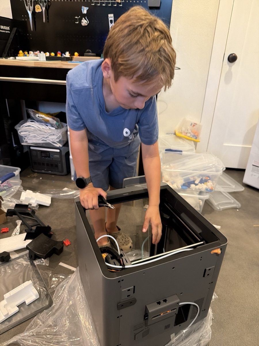 Joseph, eight, leaning into a new 3D printer with a pair of snips, fitting the filament tube, packaging still on the floor