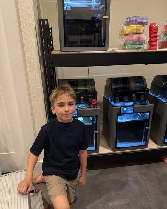 Joseph in front of a row of the Lab's real 3D printers, with filament spools on the shelf above