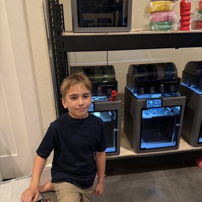 Joseph kneeling in front of a shelf of 3D printers at the Printer Ranch in Austin